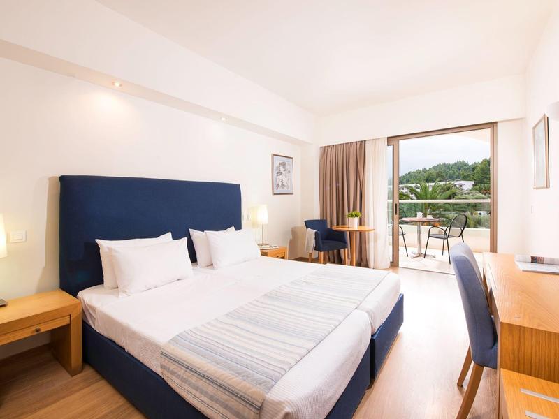 Habitación de hotel luminosa con cama doble, escritorio, silla y balcón con vista verde.