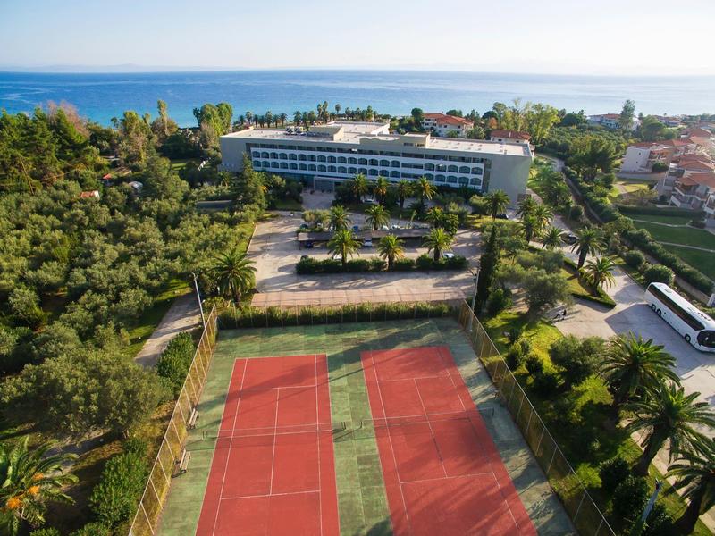Hotel con cancha de tenis rodeado de vegetación y vista al mar en el horizonte.