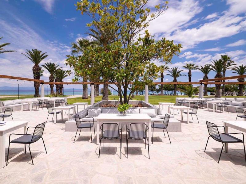 Terraza al aire libre con mesas y sillas, vistas al mar y palmeras bajo cielo azul con nubes.