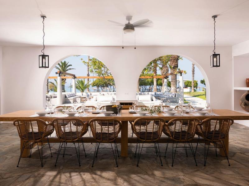Gran mesa de comedor con sillas en un espacio abierto con arcos y vista a palmeras afuera.