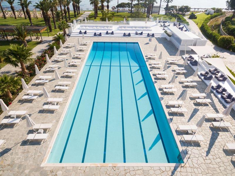 Piscina exterior rectangular con tumbonas y palmeras en un hotel junto al mar.