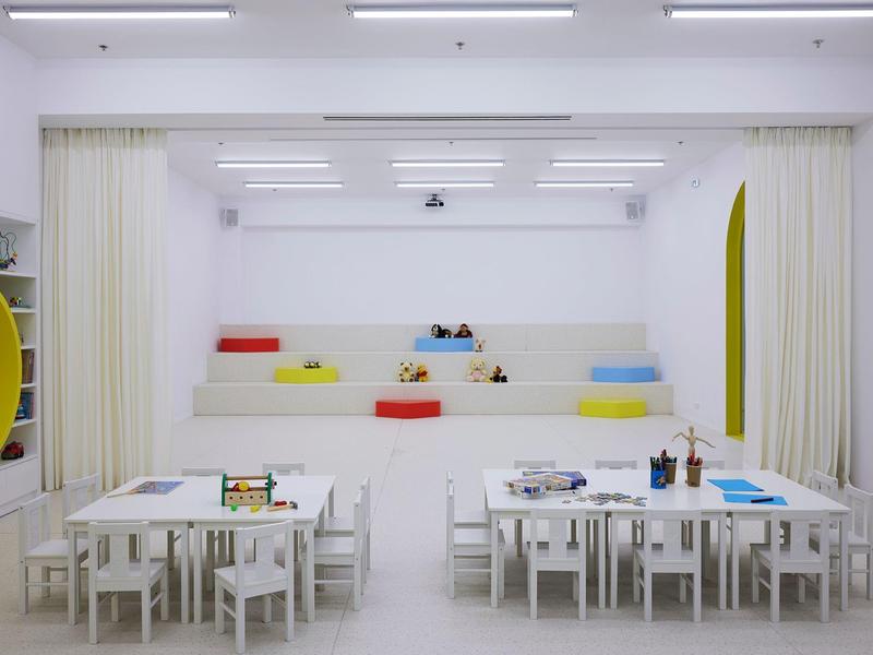 Espacio de juego infantil moderno y blanco con pequeñas mesas y cojines de colores en la pared.