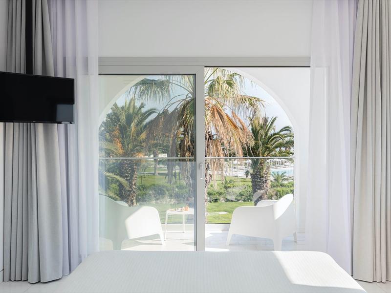 Habitación de hotel luminosa con vista a la terraza y palmeras a través de ventanas con cortinas blancas.