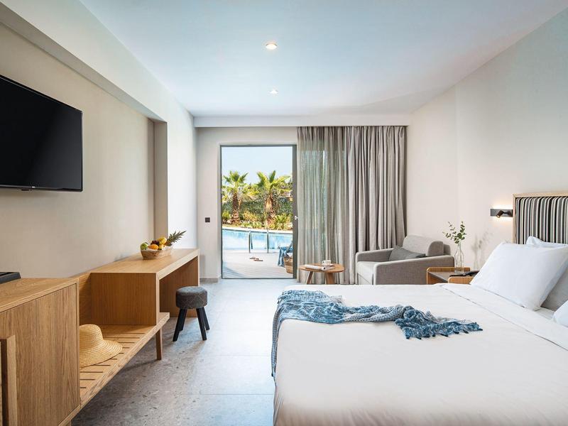 Habitación de hotel luminosa con cama grande, TV, muebles de madera y vista a la piscina con palmeras.