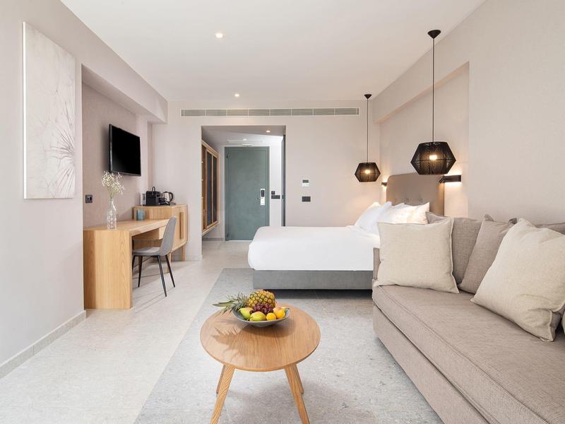 Habitación de hotel luminosa y moderna con cama, sofá, mesa pequeña y zona de escritorio.