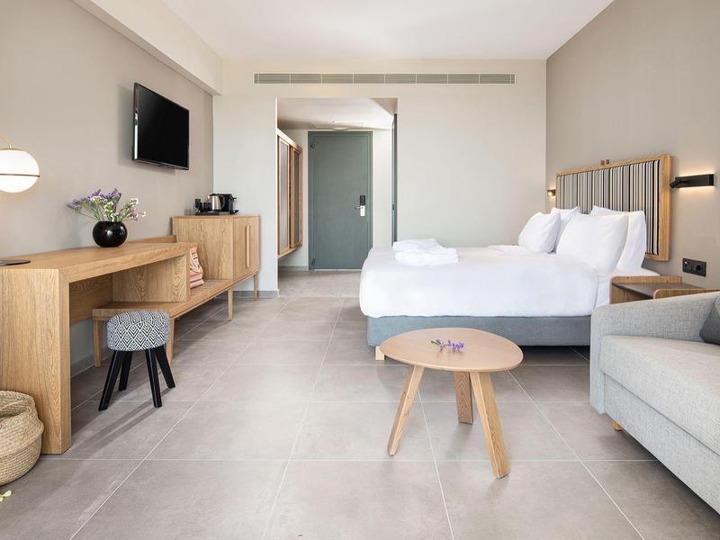 Habitación de hotel luminosa con dos camas, sofá, TV y mobiliario moderno de madera.