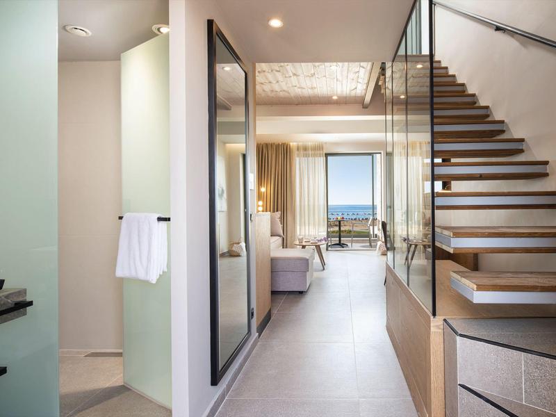 Corredor moderno de hotel con escalera de madera y vista al mar a través de grandes ventanas al fondo.