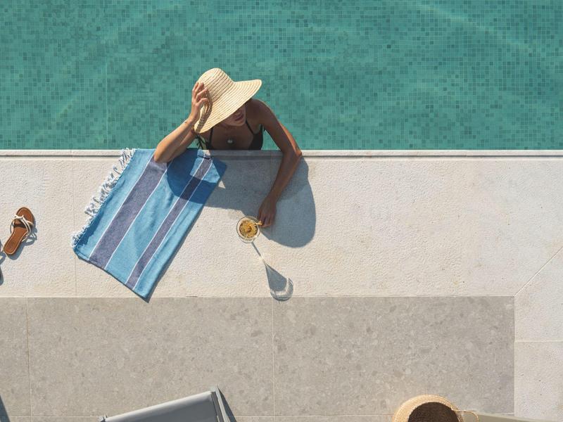 Persona con sombrero relajándose al borde de la piscina en una toalla junto a bebidas.
