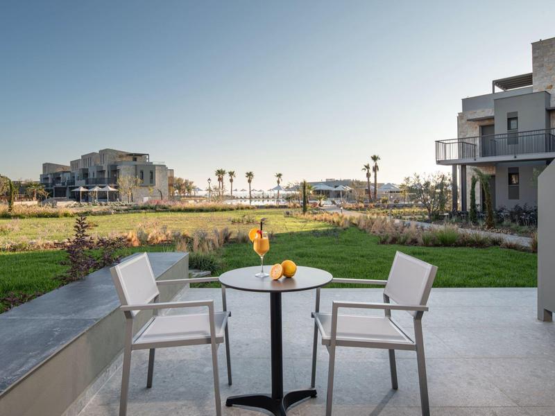 Terraza con dos sillas y una mesa con frutas, vista a campo verde y edificios al fondo.