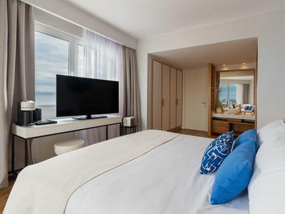 Modern hotel room with large bed, TV, wardrobe, and bathroom with sink and mirror.