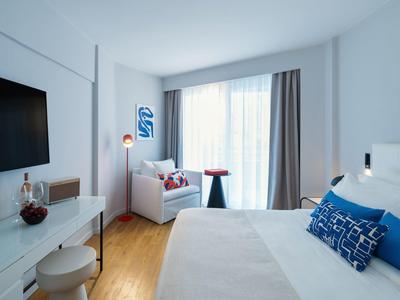 Modern hotel room with large bed, armchair, desk, and TV.