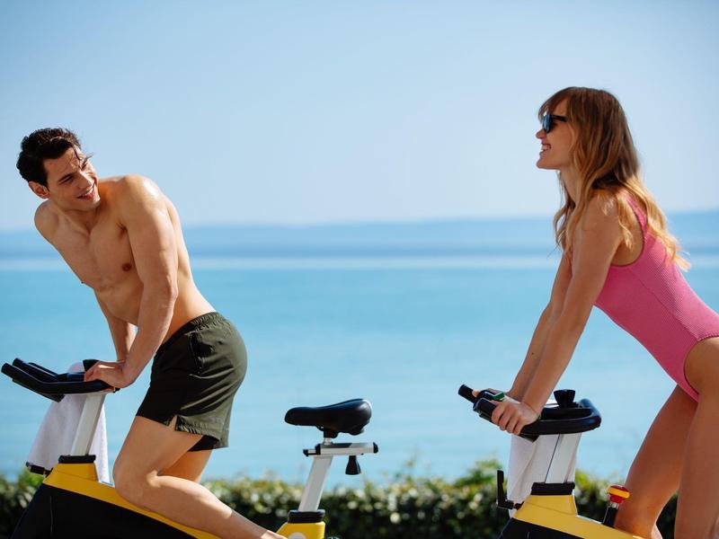 Man and woman exercising on stationary bikes by the seaside with clear blue sky.