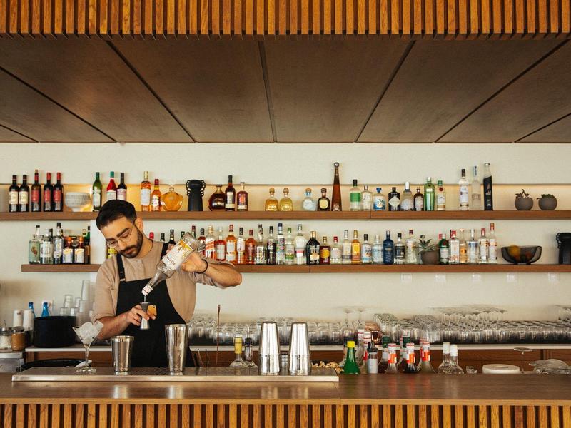 Bartender wearing mask preparing a cocktail behind a wooden bar with bottles on shelves.