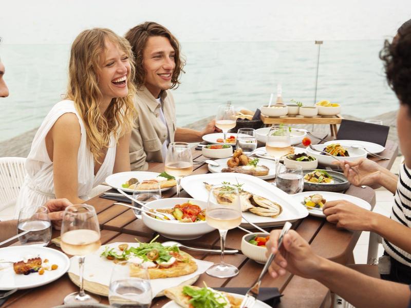 Group enjoying a meal together outdoors by the water, with plates of diverse food on the table.