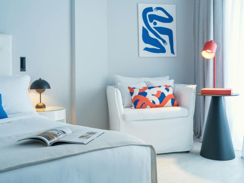 Cozy hotel room with white bed and armchair, decorative pillow, and abstract wall art.