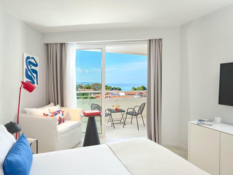 Bright hotel room with balcony, sea view, cozy couch, and modern TV.