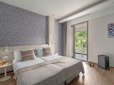 Bright hotel room with double bed, gray headboard, balcony, and view of green hills.