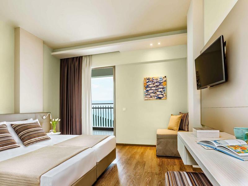 Modern hotel room with double bed, sofa, flat-screen TV, and balcony view.