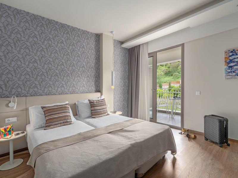 Bright hotel room with double bed, gray headboard, balcony, and view of green hills.