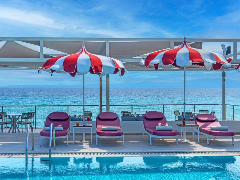 Pool area with umbrellas and lounge chairs overlooking turquoise sea and clear sky.