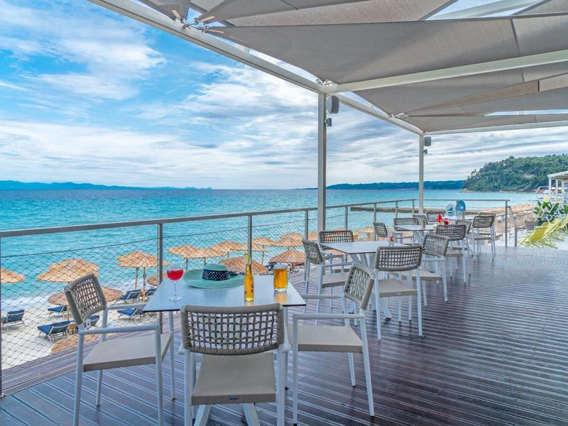 Restaurant terrace with tables and chairs by the sea under a sunshade.