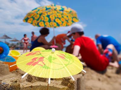 Bunter Sonnenschirm im Sand, verschwommene Personen und weitere Sonnenschirme im Hintergrund.