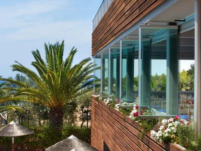 Moderner Holzbau mit großen Glasfenstern, Palmen und blühenden Pflanzen, blauer Himmel im Hintergrund.