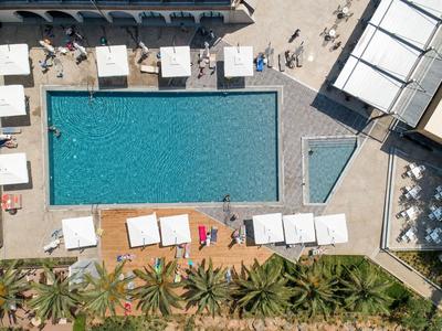 Blick von oben auf rechteckigen Pool mit Liegestühlen und Palmen neben einem Hotel.