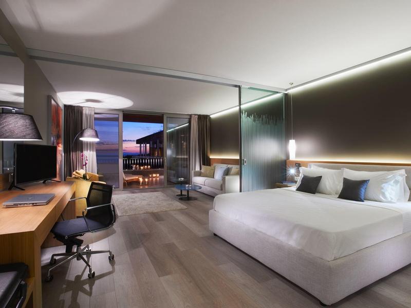 Modernes Hotelzimmer mit großem Bett, Schreibtisch, Stuhl und Glaswand zum Balkon bei Dämmerung.