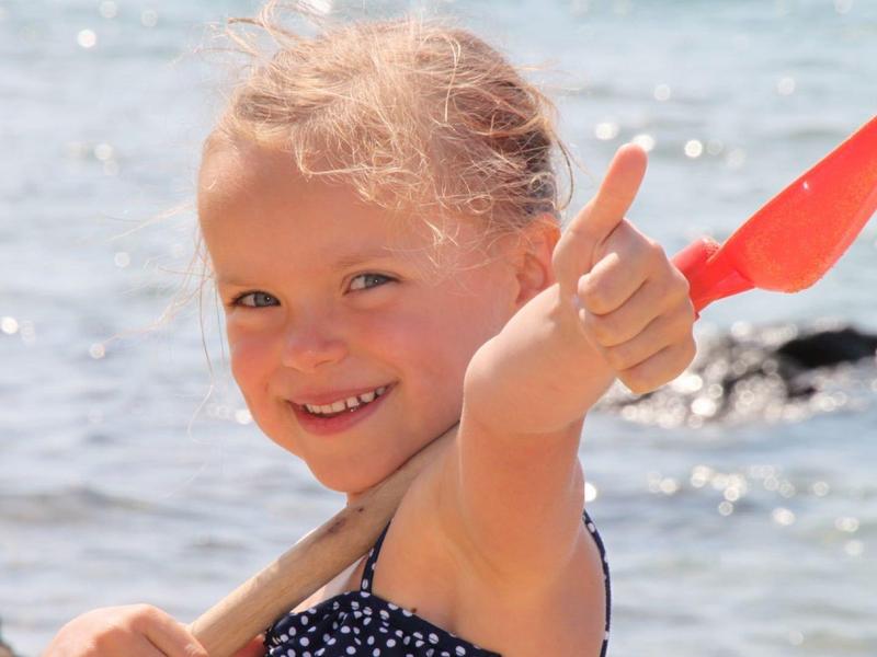 Ein lächelndes Mädchen am Wasser zeigt einen Daumen hoch, hält eine rote Sandschaufel.