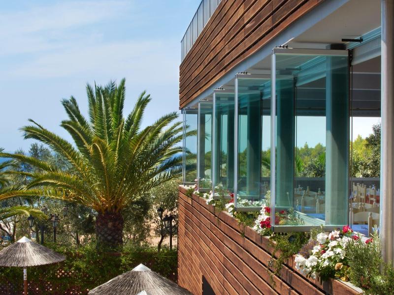 Moderner Holzbau mit großen Glasfenstern, Palmen und blühenden Pflanzen, blauer Himmel im Hintergrund.
