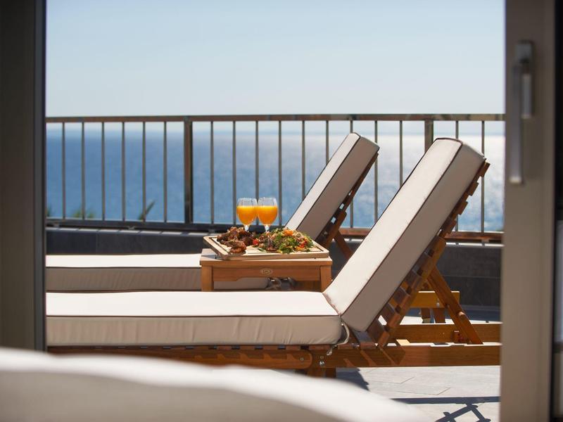 Zwei bequeme Liegestühle mit Getränken und Snacks auf Balkon mit Meerblick bei klarem Himmel.