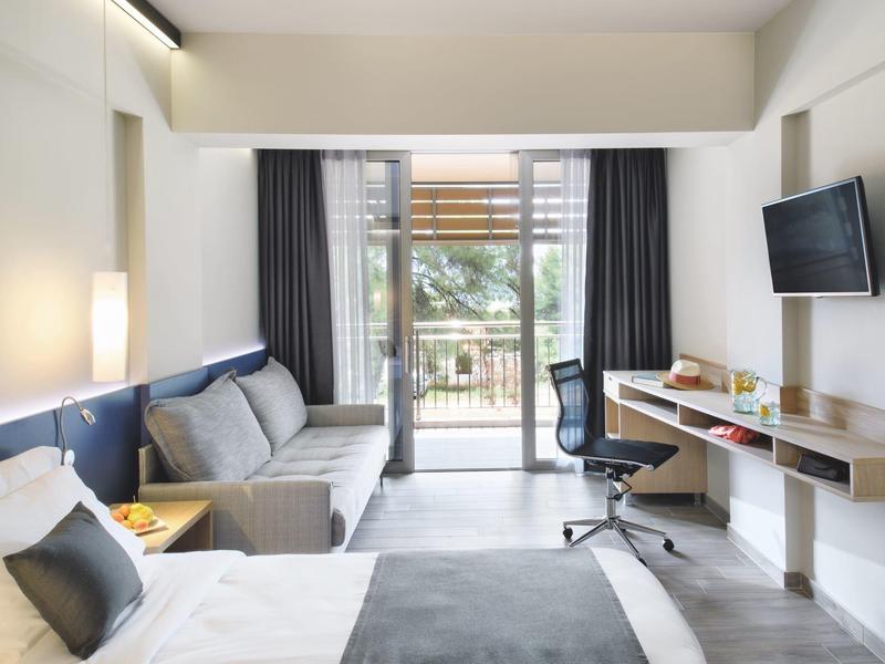 Modernes Hotelzimmer mit Bett, Sofa, Schreibtisch und Balkon mit Blick ins Grüne.