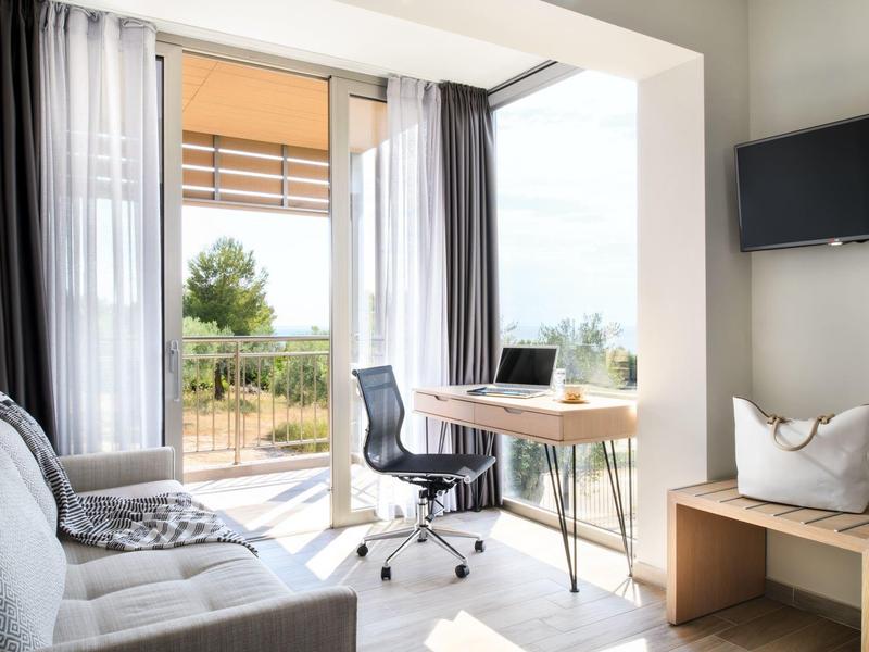 Helles Wohnzimmer mit Sofa, modernem Schreibtisch, Drehstuhl und Zugang zu Balkon mit grünem Ausblick.