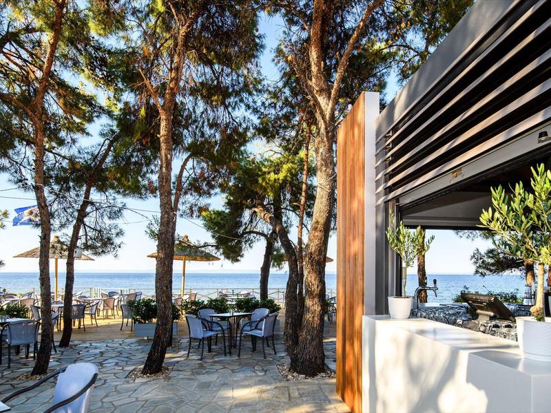 Freiluftcafé unter Bäumen mit Blick auf das Meer und Tische mit Stühlen auf hellem Steinboden.