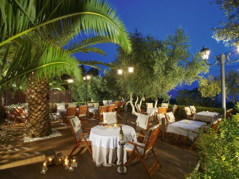 Abendliche Terrasse mit Palmbaum, Tischen mit weißen Tüchern, Holzmöbeln und Lichterketten.