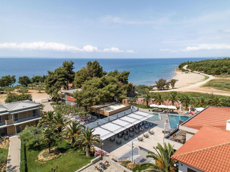 Blick auf ein Gelände mit Häusern, Pool, Palmen und Blick auf das Meer und den Strand im Hintergrund.