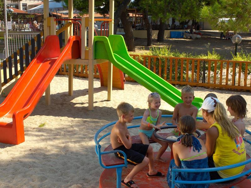 Children sit on a carousel at a sandy playground with colorful slides in the background.
