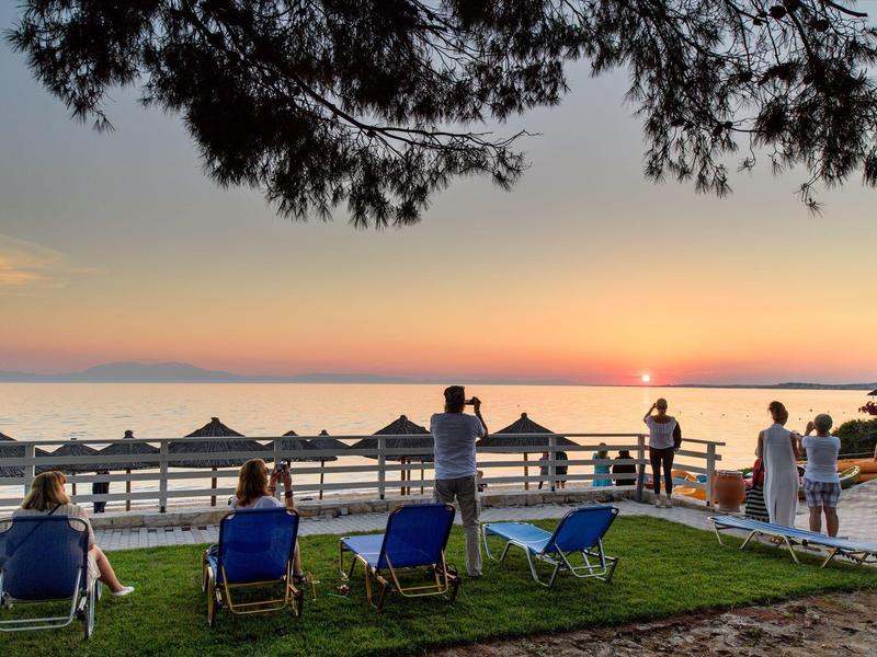 Sunset by the sea with lounge chairs and people by the water's edge.