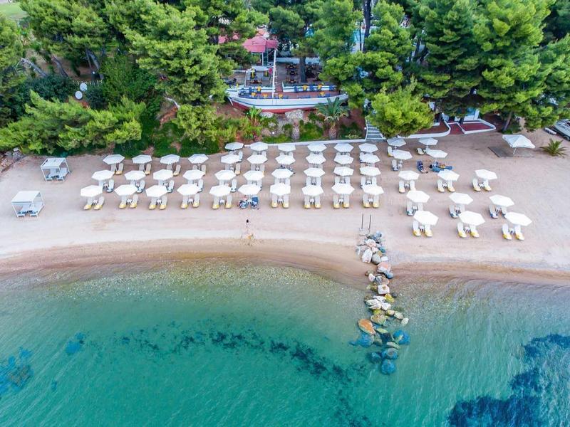 Strand mit weißen Liegestühlen und Sonnenschirmen, türkisfarbenem Wasser und dichter grüner Vegetation.