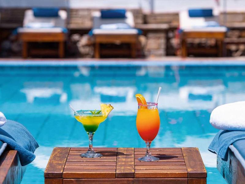 Ein Pool mit blauer Wasserfläche, umgeben von Liegestühlen, zwei bunte Cocktails auf einem Holztisch.