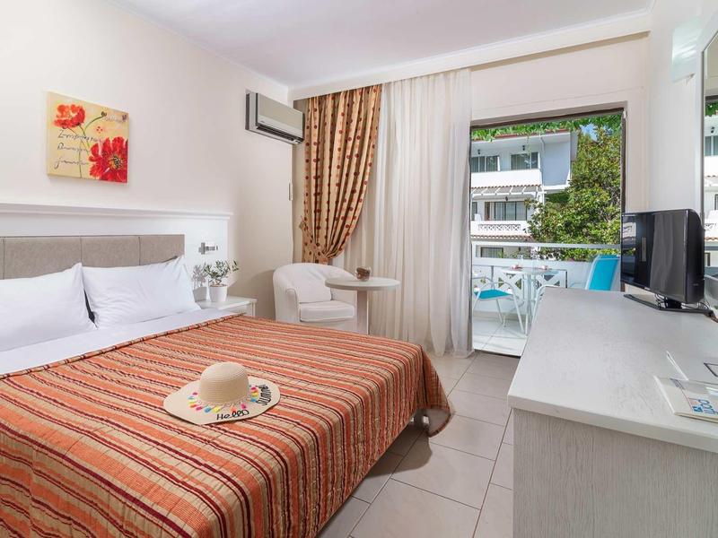 Helles Hotelzimmer mit Doppelbett, gestreifter Decke, kleinem Balkon und heller Einrichtung.