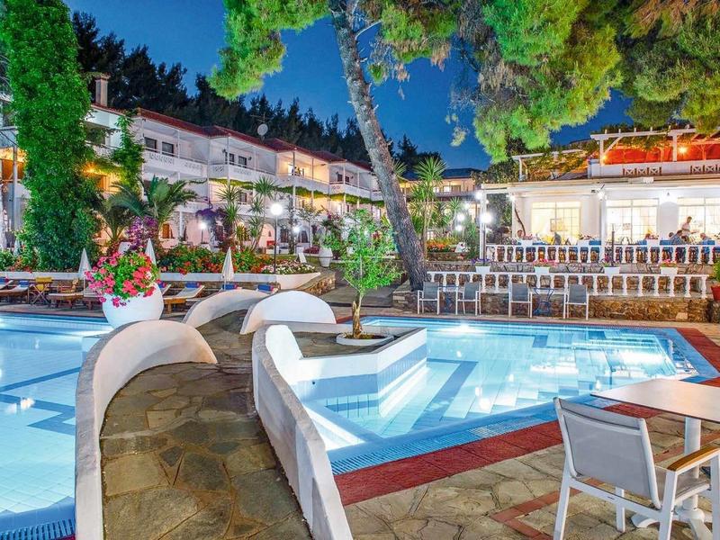 Beleuchteter Poolbereich mit bepflanzten Bäumen, Terrasse und Tischen vor einem Restaurant bei Nacht.