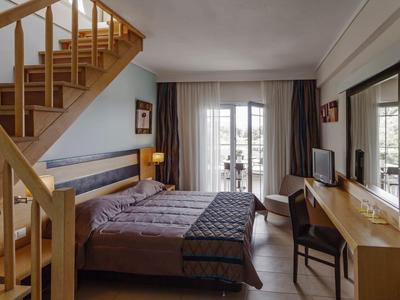 Příjemný hotelový pokoj se dvěma lůžky, dřevěným schodištěm, psacím stolem, televizí a balkonovými dveřmi se závěsy.