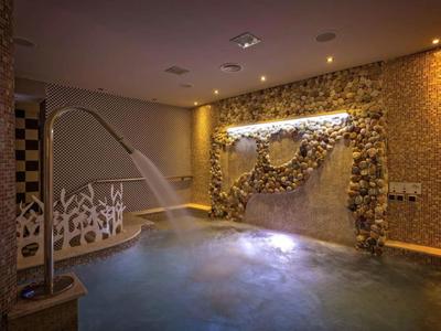 Vnitřní lázeňský bazén s tekoucím vodním prvkem a teplým osvětlením kamenné stěny.