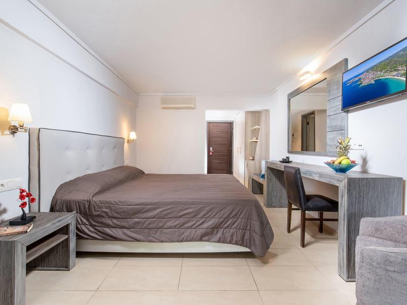 Světý hotelový pokoj s postelí, psacím stolem, židlí, televizí na stěně a měkkým osvětlením.