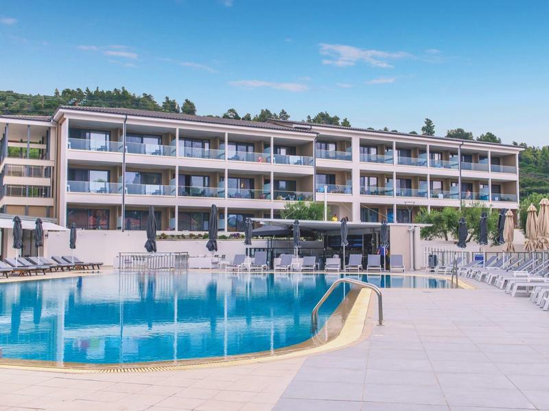 Complexe hôtelier moderne avec grande piscine, terrasse ensoleillée et chaises longues devant colline boisée.