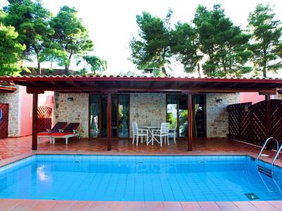 Helles Haus mit rotem Dach, großem Pool und Terrasse, umgeben von grünen Bäumen.