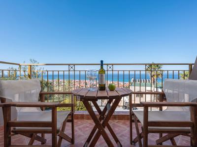 Balkon mit zwei Holzstühlen, kleinem Tisch, Pflanzen und Meerblick unter blauem Himmel.