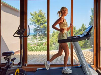 Woman running on treadmill in bright gym with view of garden and sea.